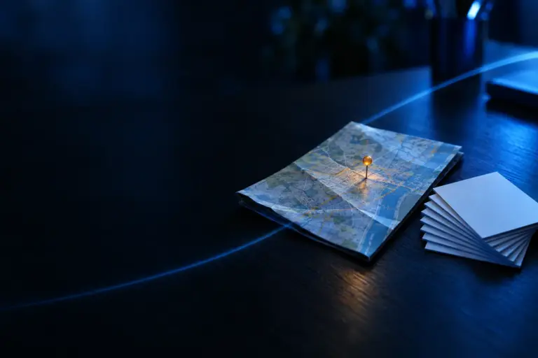 Folded city map on dark surface with orange location pin and stack of blank business cards nearby under blue lighting